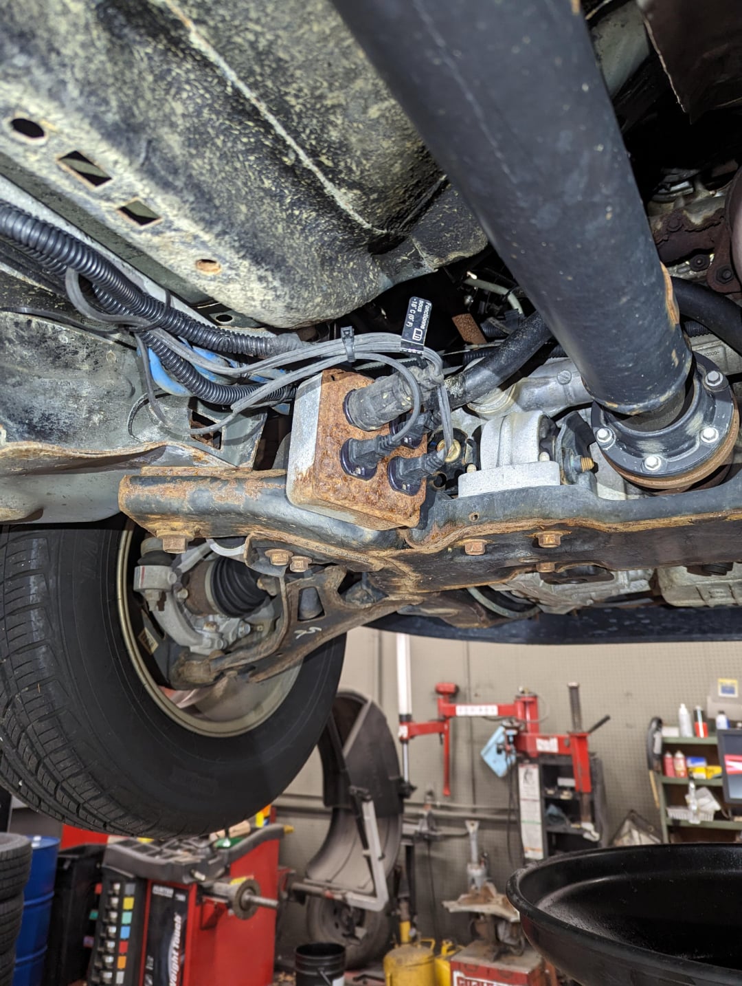 View from beneath a vehicle showing the suspension, wheel, drive shaft, and rusted components of the chassis, with automotive repair equipment visible in the background.