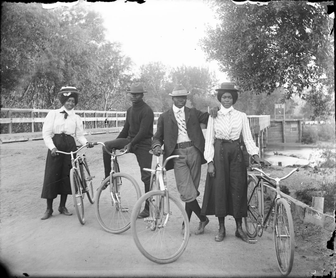 Four people in early 20th-century clothing stand outdoors with bicycles on a dirt road near a wooden bridge, trees in the background. Two women and two men are posing and smiling for the camera.