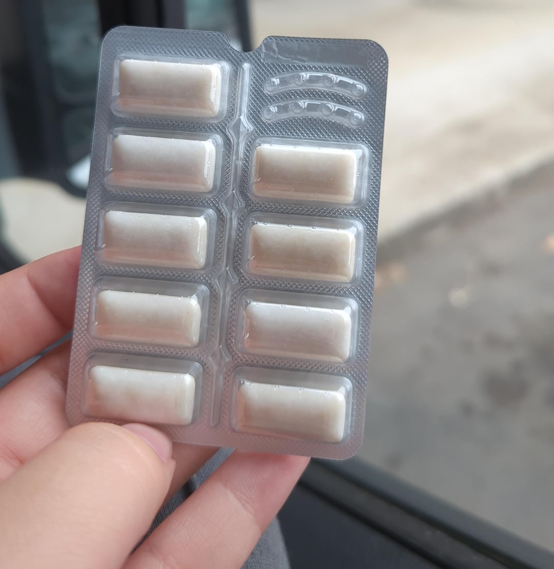 A hand holding a blister pack containing ten white, rectangular tablets, with an outdoor background visible in soft focus.