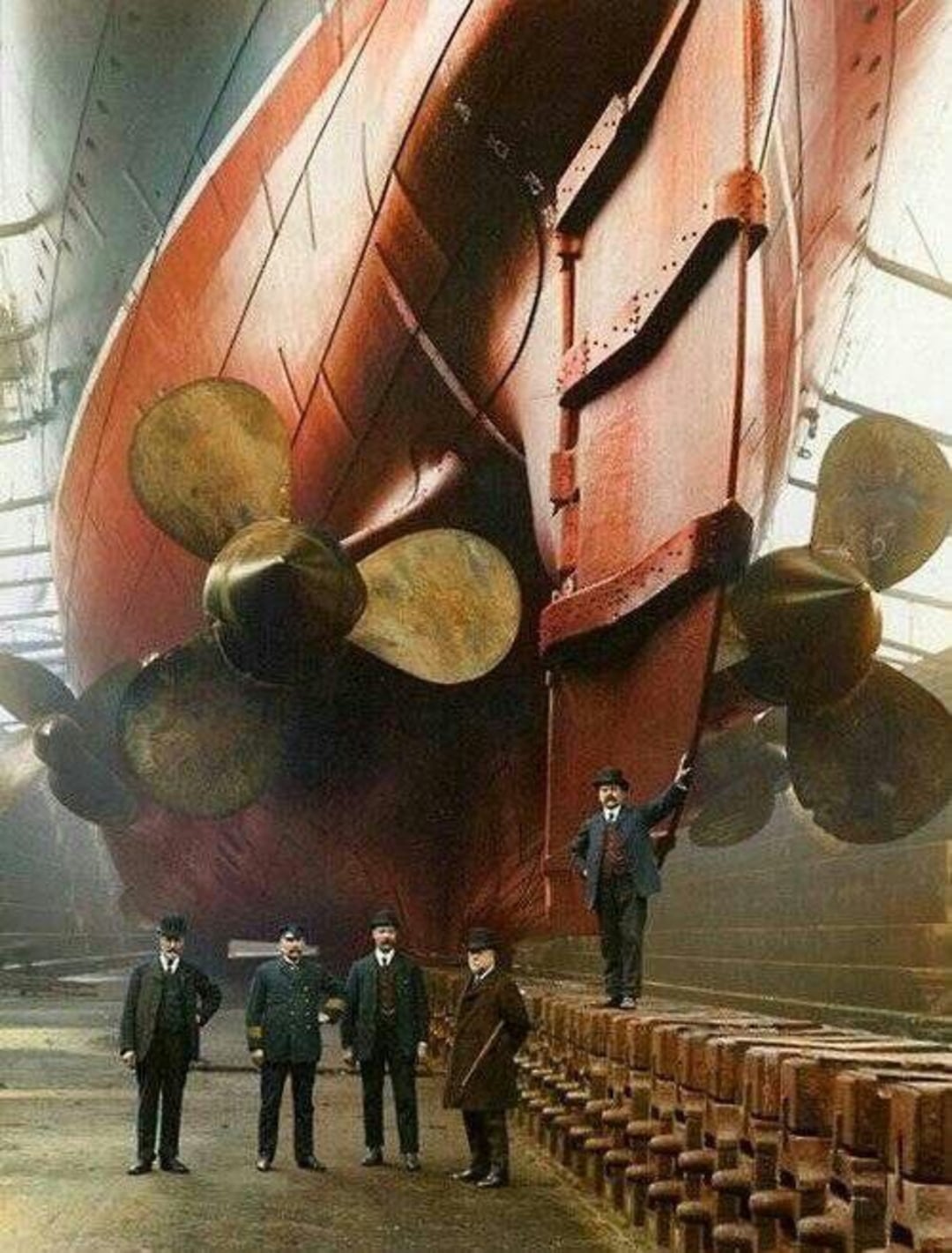 Five men in early 20th-century attire stand beneath the massive propellers and rudder of a large ship in a dry dock, highlighting the ship's enormous scale.