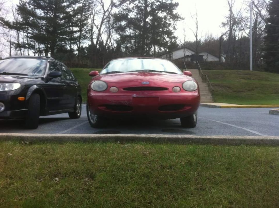 A red car is parked in a lot next to a black car, with grass in the foreground and trees, a hill, and a house in the background on an overcast day.