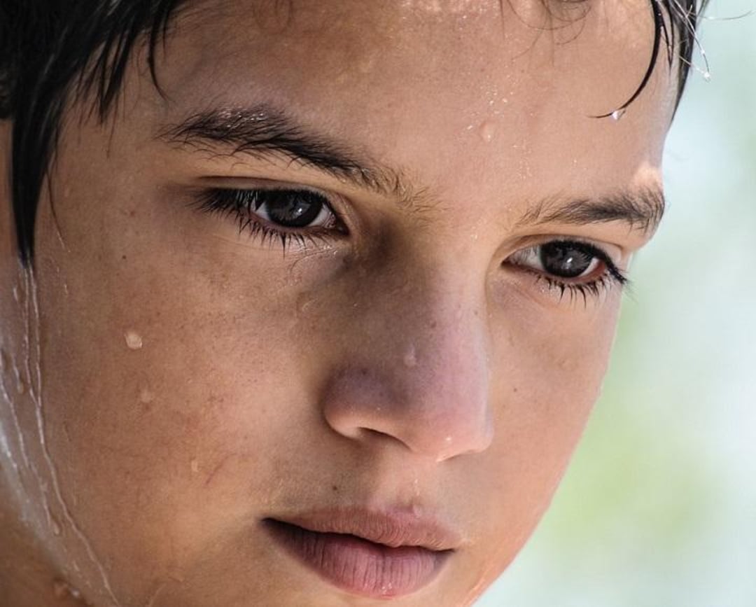 Close-up of a young person's face with wet skin and water droplets, gazing downward with a thoughtful expression. The background is softly blurred in light colors.