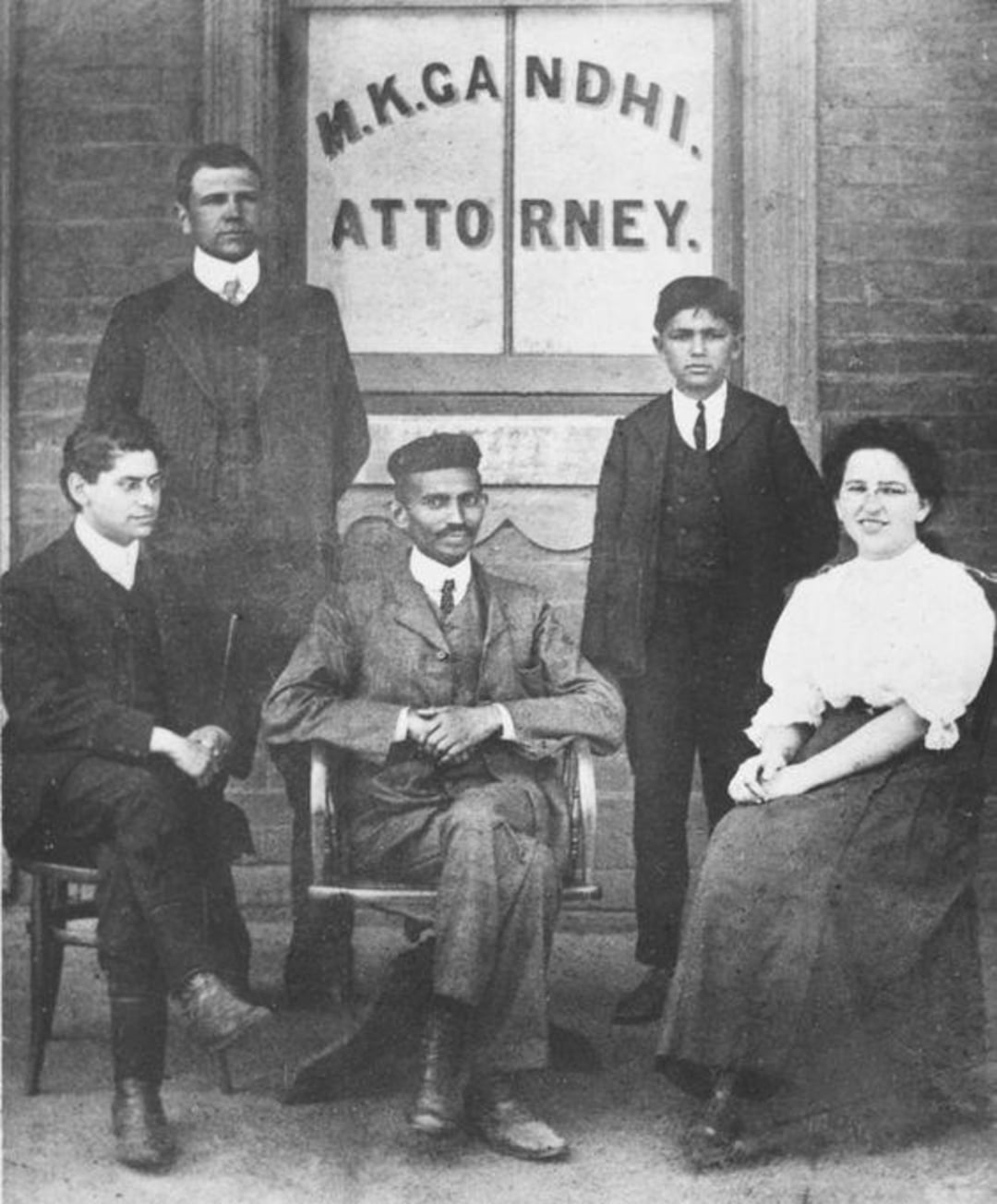 Five people pose in front of a building window labeled “M.K. Gandhi. Attorney.” Four men or boys, one seated woman, all in early 20th-century Western attire, with the central man seated and others standing or sitting around him.