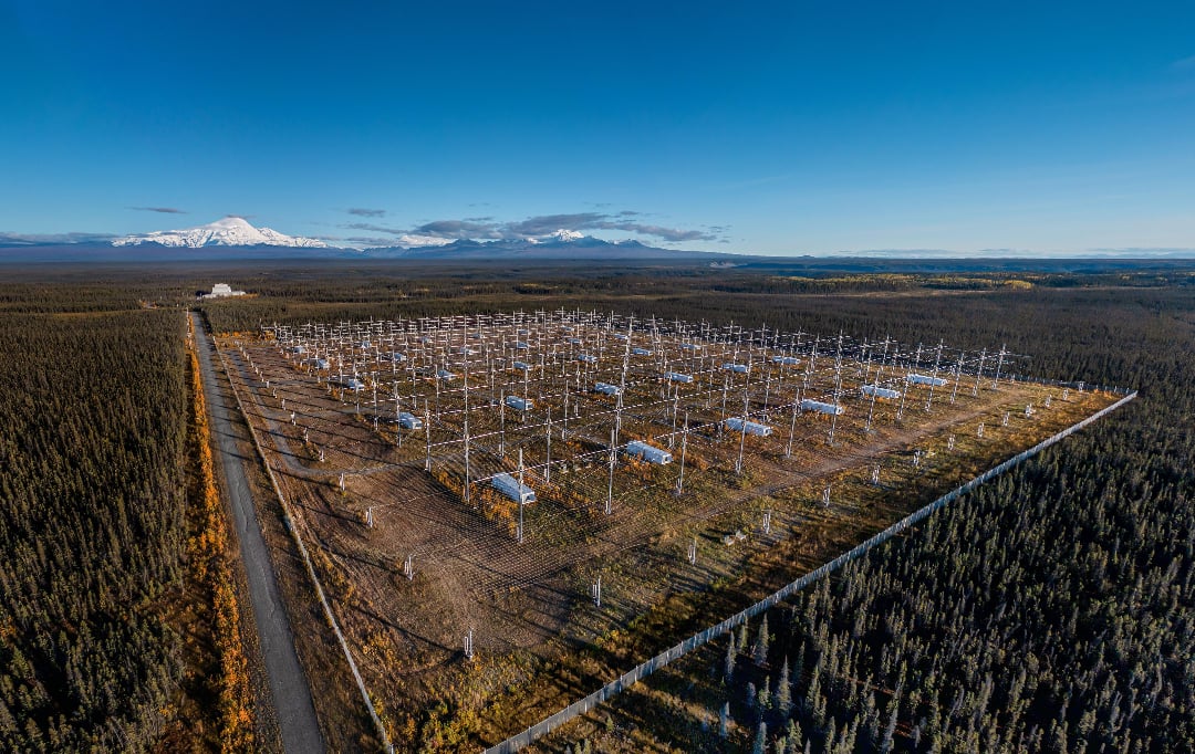 Aerial view of a large research facility with rows of tall metal antennas in a clearing, surrounded by dense forest. Snow-capped mountains are visible in the distance under a clear blue sky.