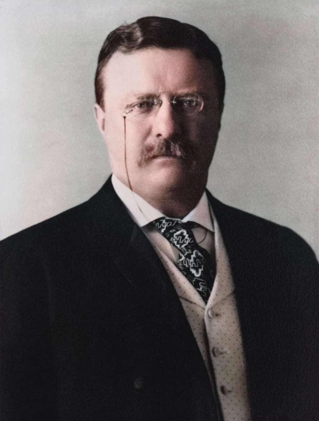 A man with a mustache wearing glasses, a dark suit, a white shirt, a patterned tie, and a light-colored vest, poses for a formal portrait against a plain background.