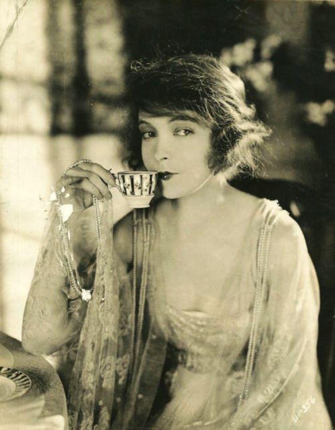 A woman in vintage attire and sheer shawl sips tea from a patterned teacup, gazing directly at the camera in a softly lit, elegant setting.