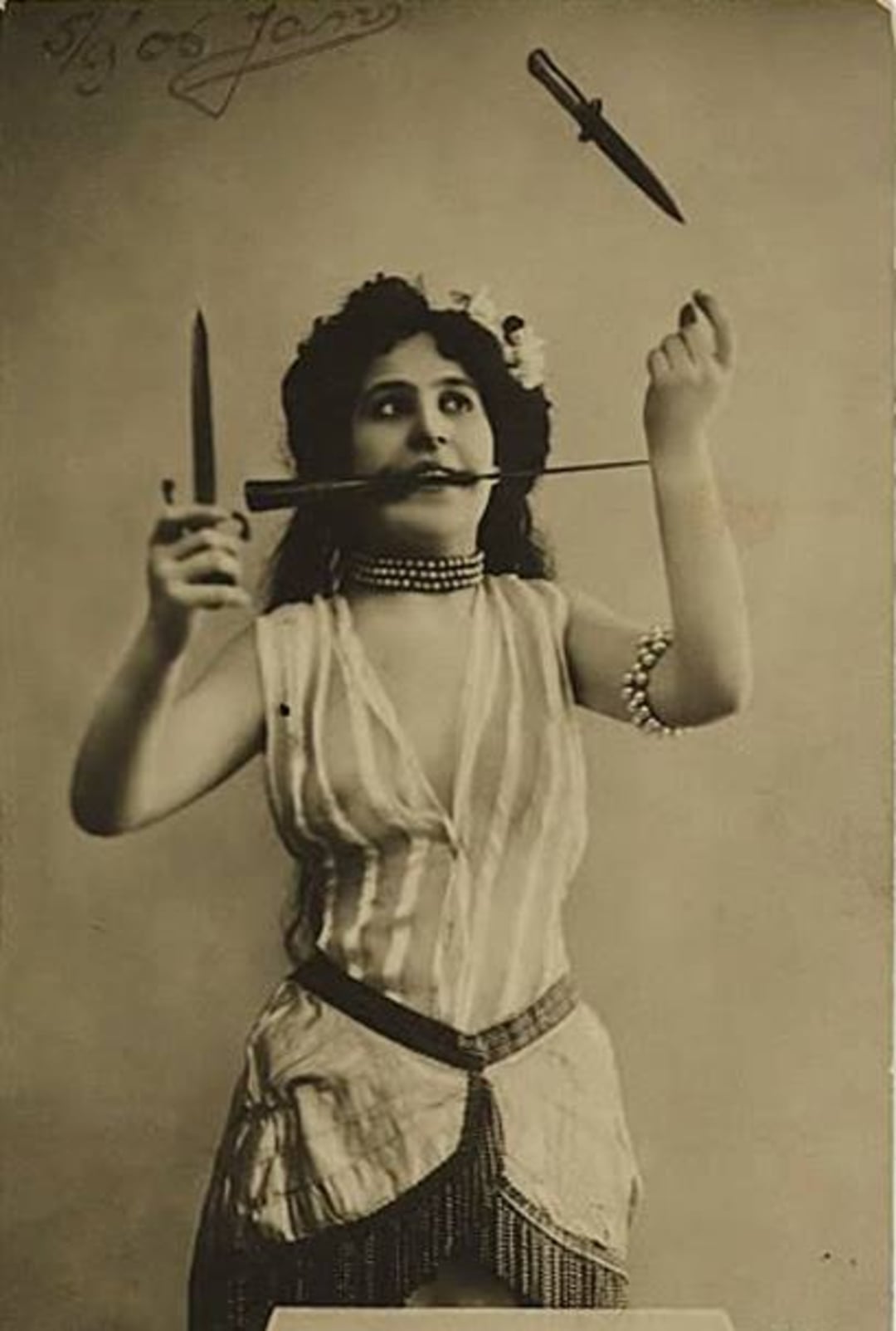 A woman in vintage attire balances a dagger in her mouth, holds one in her hand, and tosses another into the air, performing a dramatic knife act. She has flowers in her hair and wears layered necklaces.