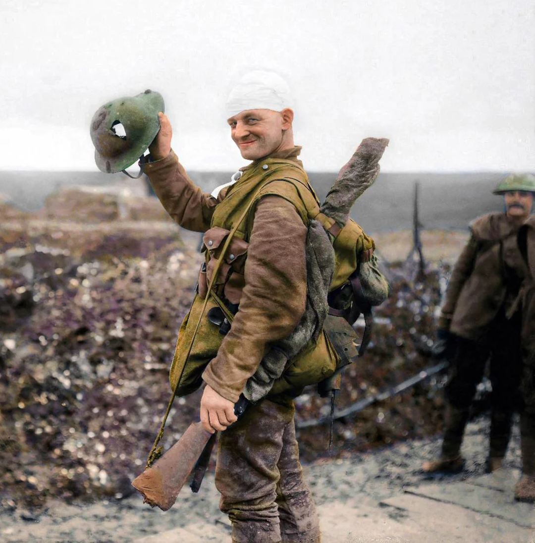 A World War I soldier with a bandaged head smiles while holding his helmet in one hand and carrying a rifle and gear on his back. Another soldier stands blurred in the background on a muddy battlefield.