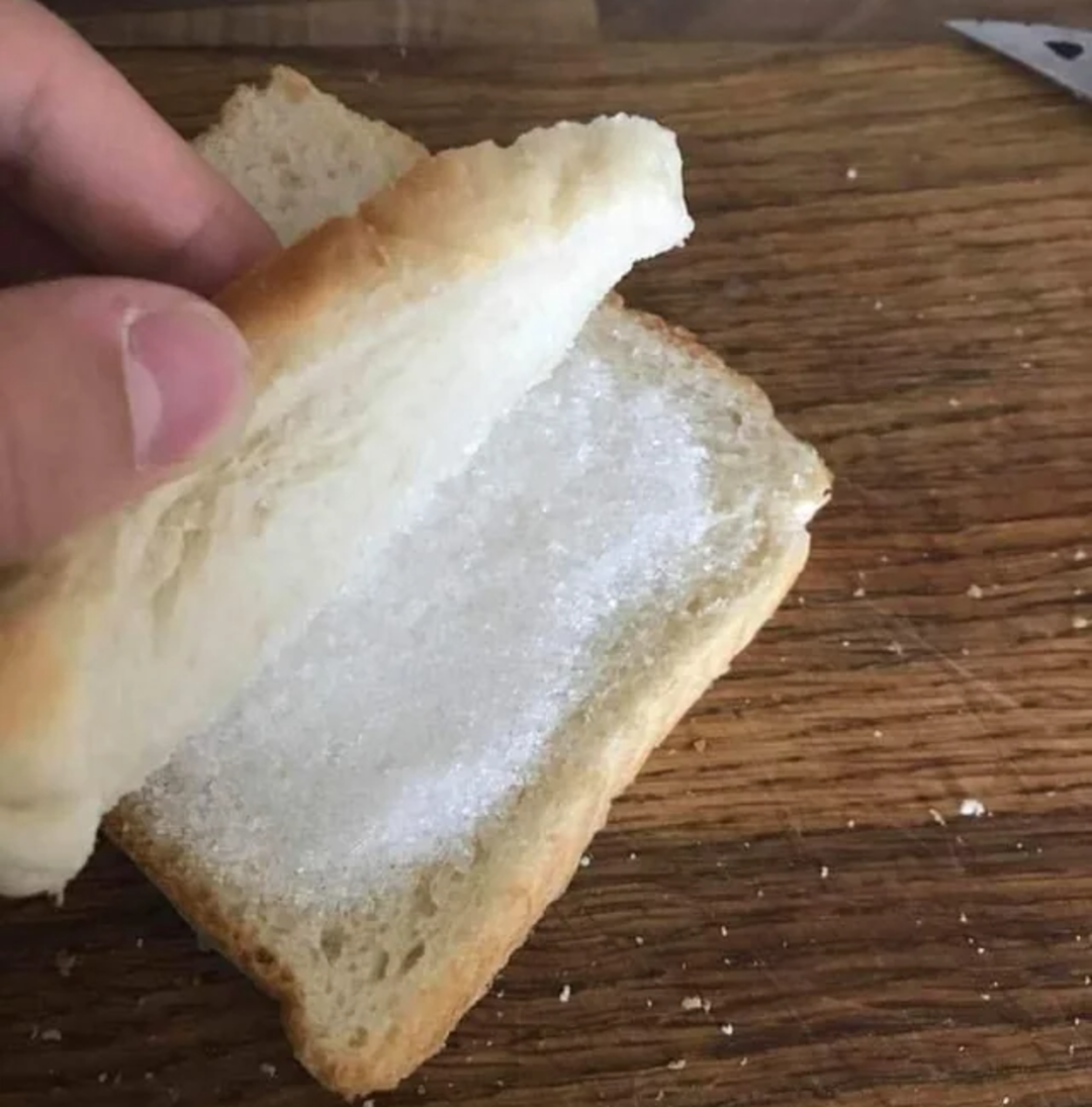 A hand lifts a slice of plain white bread, revealing another slice topped with a layer of granulated white sugar on a wooden surface.