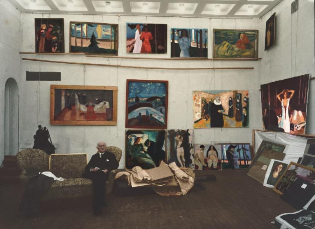 An elderly man sits on a sofa in a spacious art studio, surrounded by numerous colorful paintings on the walls and floor, with high ceilings and a wooden floor.