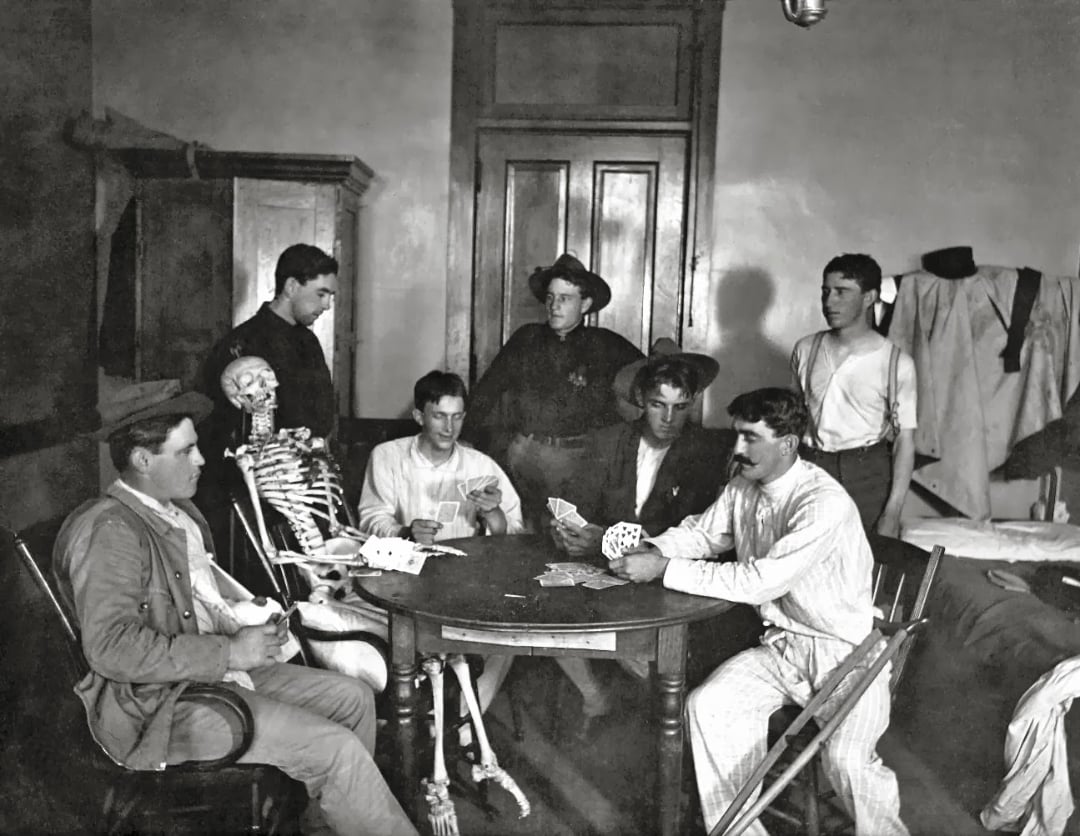 Seven men sit around a table playing cards in a vintage room; a human skeleton, dressed in clothes, is seated with them, appearing to participate in the card game.