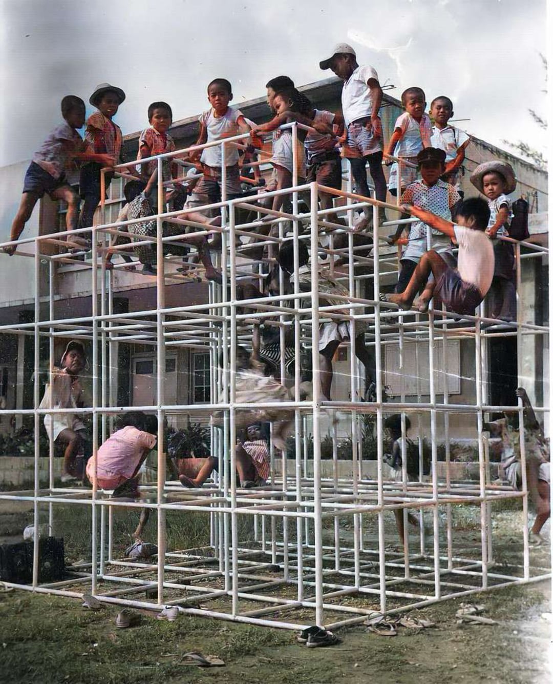 Children climb and play on a large metal jungle gym structure outdoors, with a building and grassy area in the background. The kids appear energetic and are wearing casual summer clothes.
