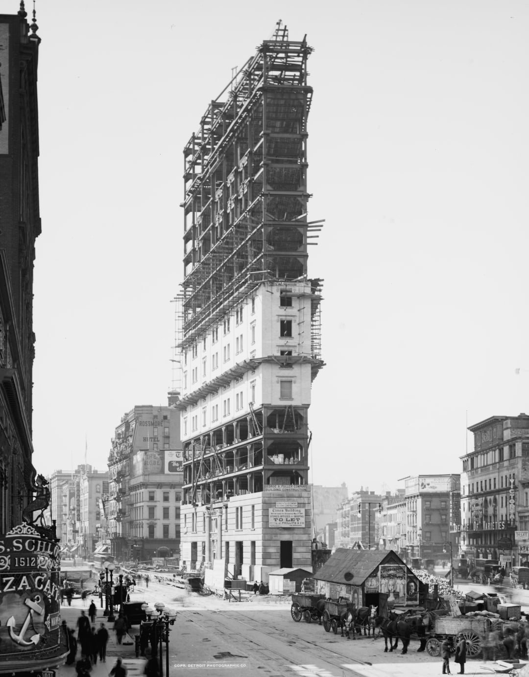 A tall building under construction stands in a busy city street, surrounded by scaffolding. People, horse-drawn carriages, and early automobiles move along the road; signs and storefronts line the street.