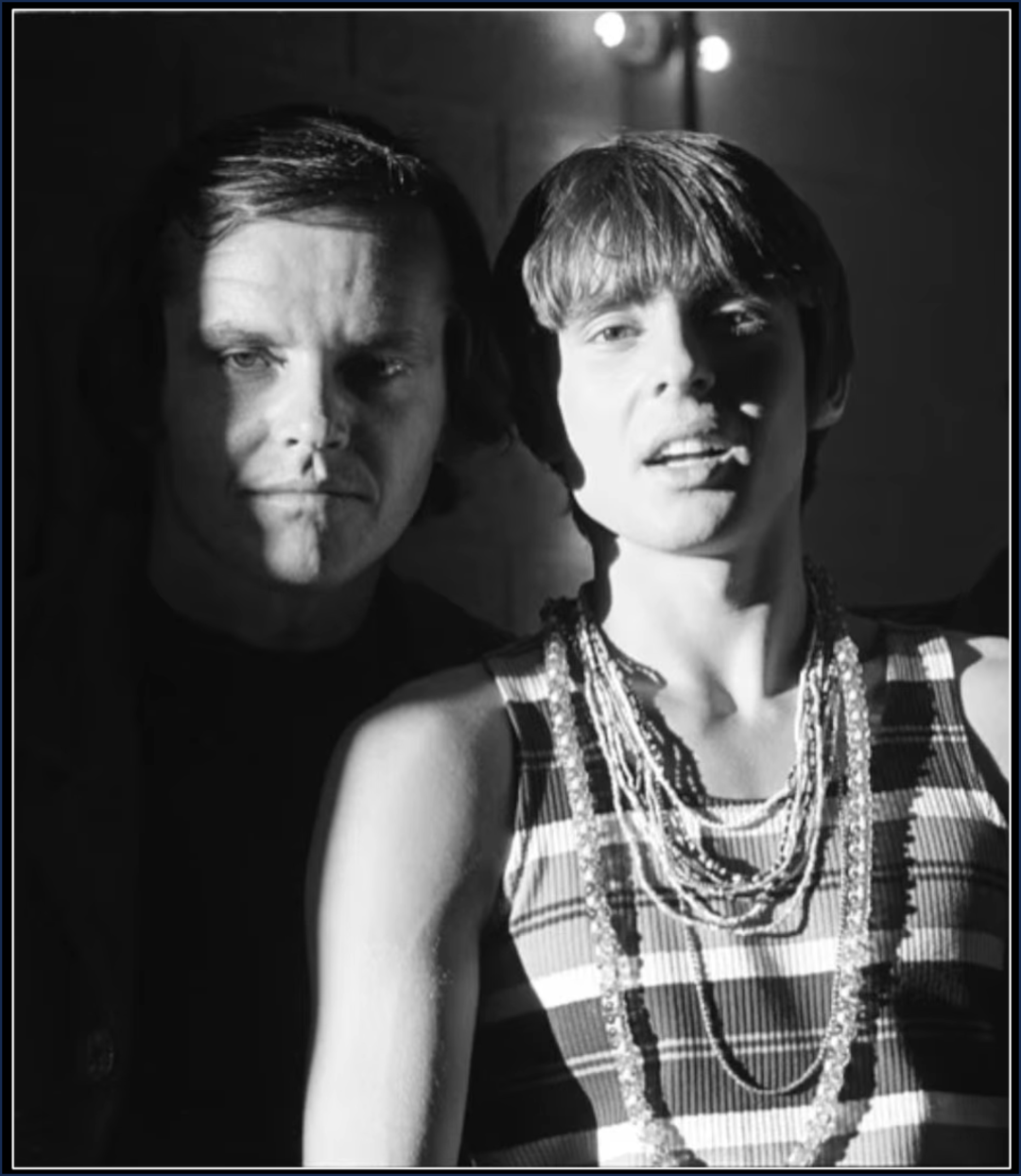 Two people stand closely together in dramatic lighting; one is in partial shadow while the other, wearing a striped top and layered necklaces, is more brightly lit. The background is dark and indistinct.