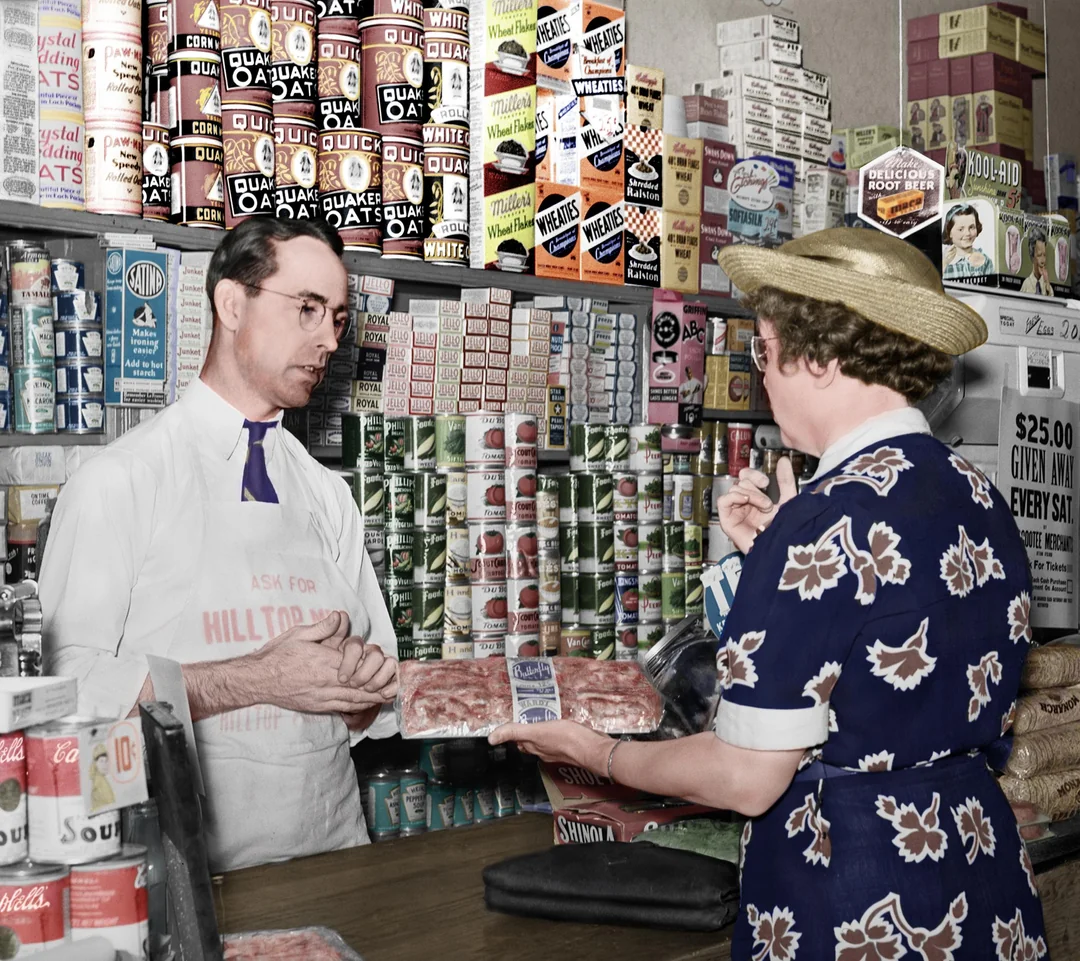 A man in a white apron and glasses stands behind a grocery store counter, talking to a woman in a floral dress and straw hat holding packaged meat, with shelves of canned and boxed goods behind them.