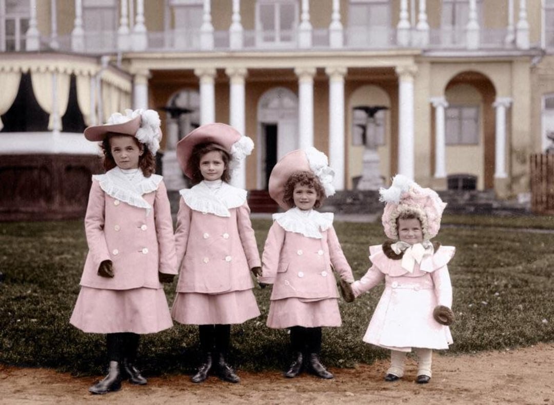 Four young girls in matching pink coats and hats stand holding hands in front of a large, elegant building with white columns and arched windows. The youngest girl is noticeably shorter than the others.