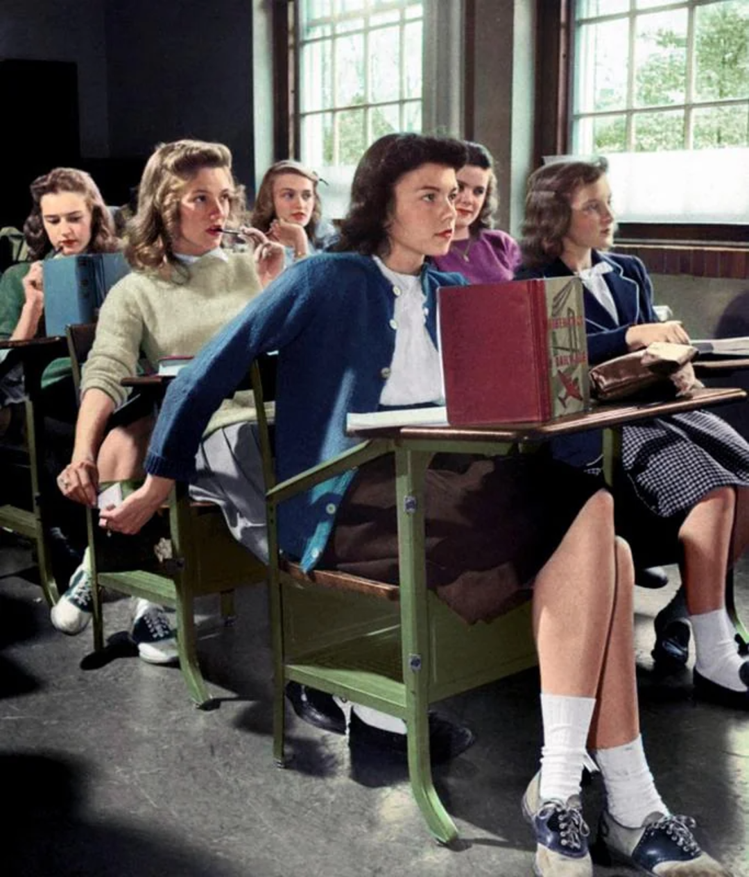 A group of teenage girls sit at their desks in a classroom, wearing 1940s-style clothing. One girl secretly passes a note to another behind their desks while others focus on the lesson. Sunlight streams through large windows.