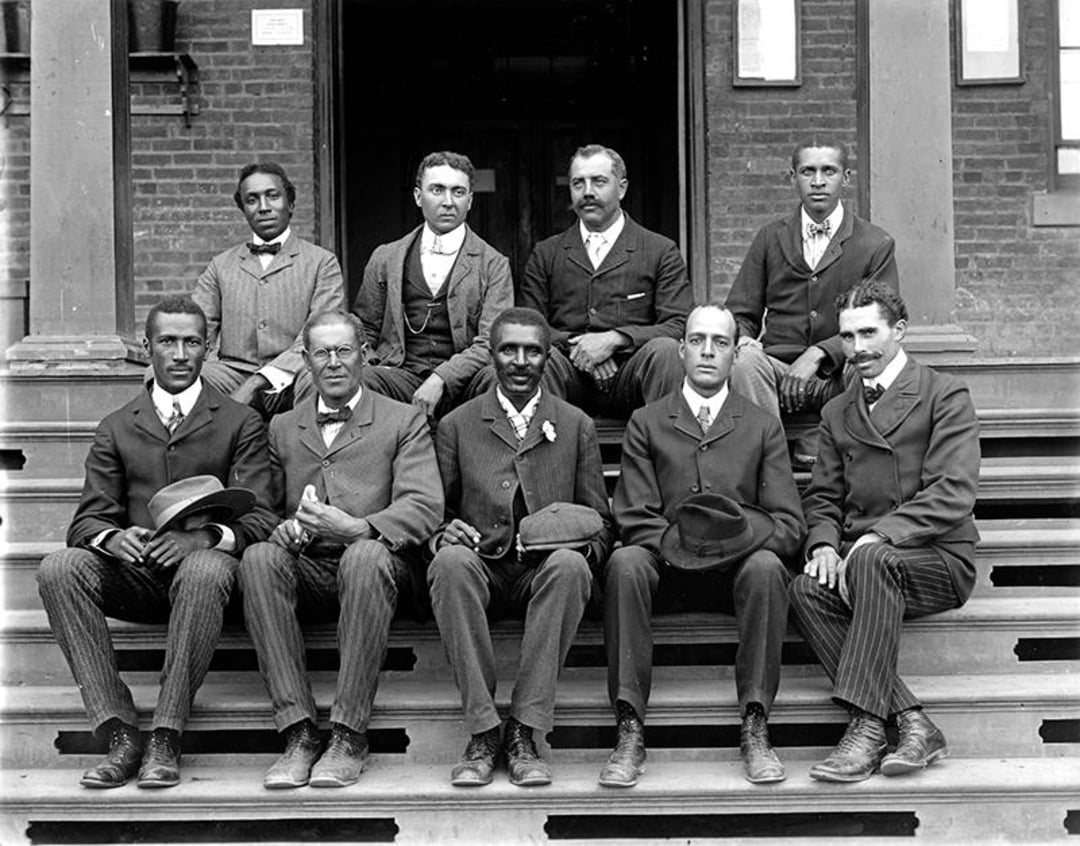 Nine men in suits pose for a formal group photo on the steps of a brick building. They are seated and standing in two rows, looking at the camera with neutral expressions.