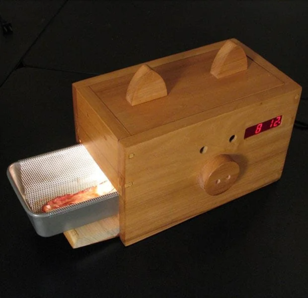 A wooden alarm clock shaped like a pig dispenses a metal tray with cooked bacon, lit from inside. The display shows the time "8:12". The design includes pig ears, eyes, and a snout on the front.