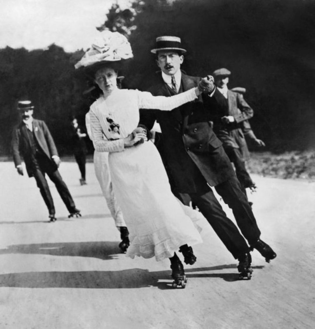 A black-and-white photo of a woman in a long white dress and wide-brimmed hat roller skating hand-in-hand with a man in a suit and boater hat, with other skaters behind them, outdoors on a paved path.