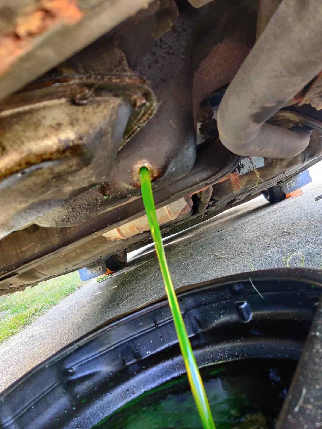 Bright green coolant flows from a vehicle's undercarriage into a black collection pan during a fluid change on a driveway.