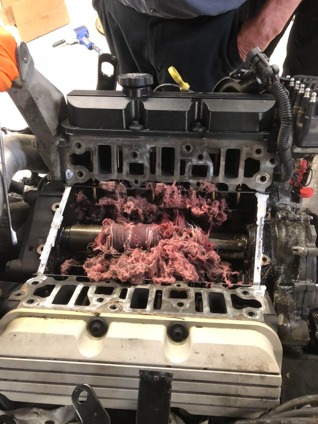 A car engine with its cover removed, exposing the camshaft tangled with shredded pink insulation-like material, while two people inspect the engine.