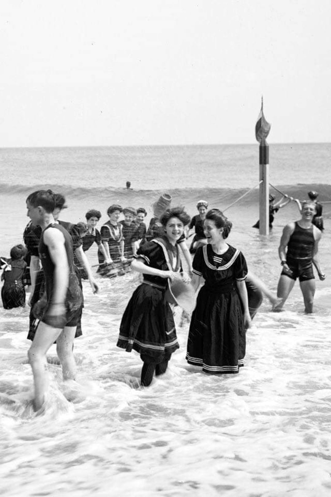 Women and men in old-fashioned swimsuits play in the shallow water at a beach. Two women in the foreground smile as waves splash around their feet. More people are seen enjoying the sea in the background. Black and white photo.