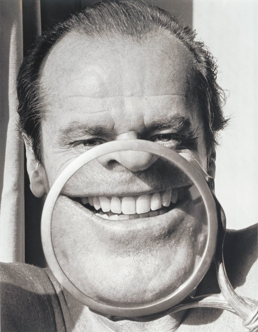 A man holds a magnifying glass in front of his mouth, making his smile appear exaggeratedly large and distorted compared to the rest of his face.