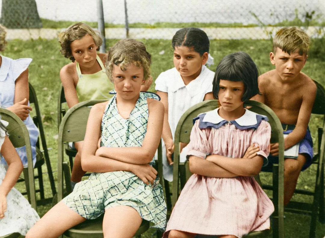 A group of children sit outdoors on folding chairs, looking serious or upset. Two girls in front sit with arms crossed. The grass and a chain-link fence are visible in the background.