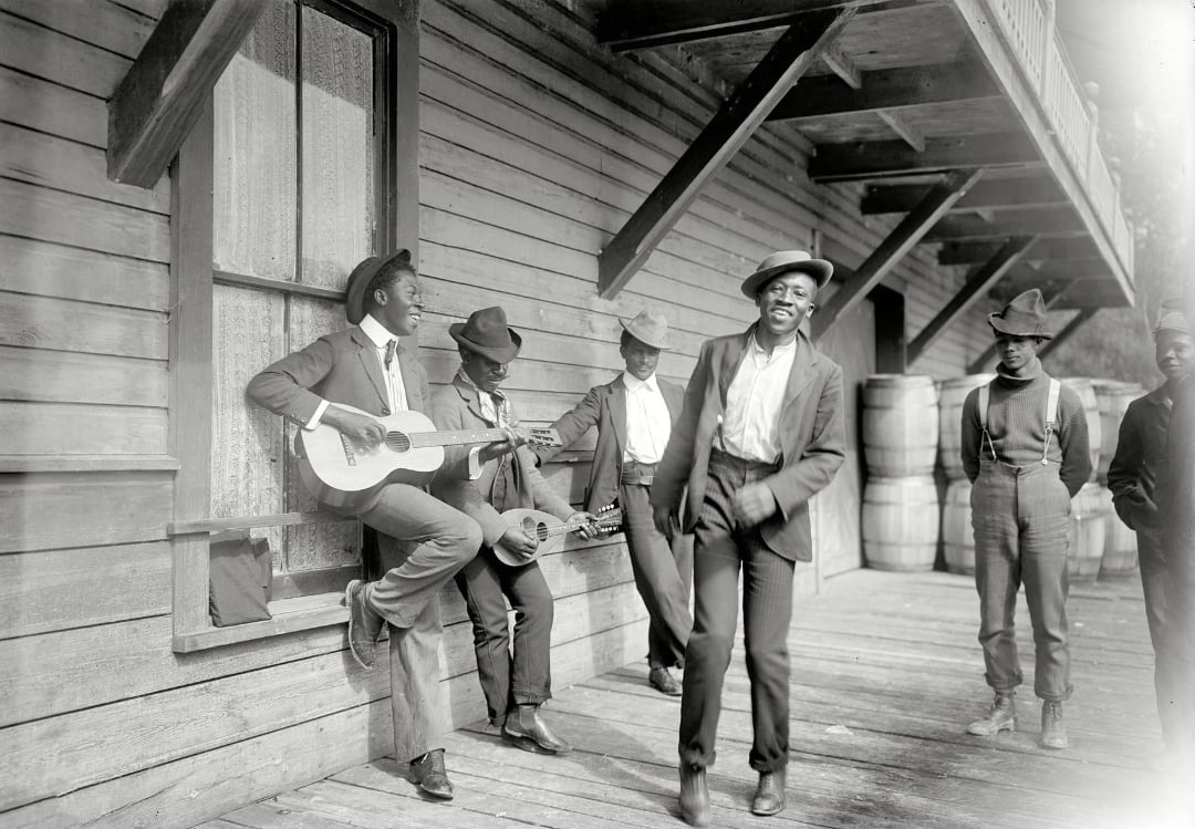 Five Black men in early 20th-century clothing stand on a wooden porch; one plays a guitar, another dances, and three watch. Barrels are stacked in the background near the wooden building.