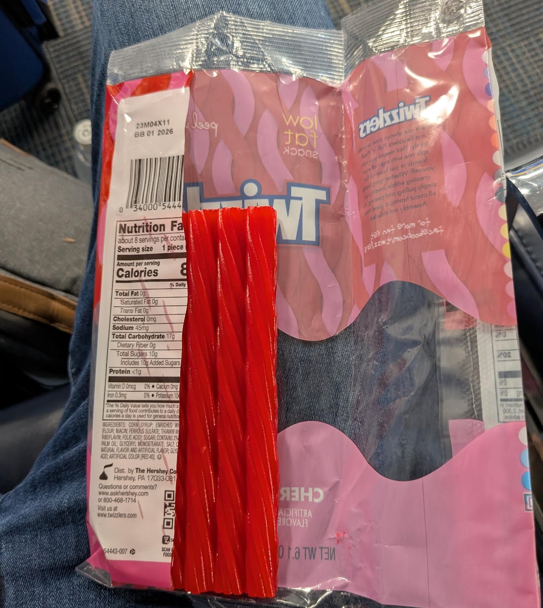A partially empty package of Twizzlers cherry candy rests on someone's lap, with a few red licorice sticks placed on top of the open bag. Nutritional information and branding are visible.