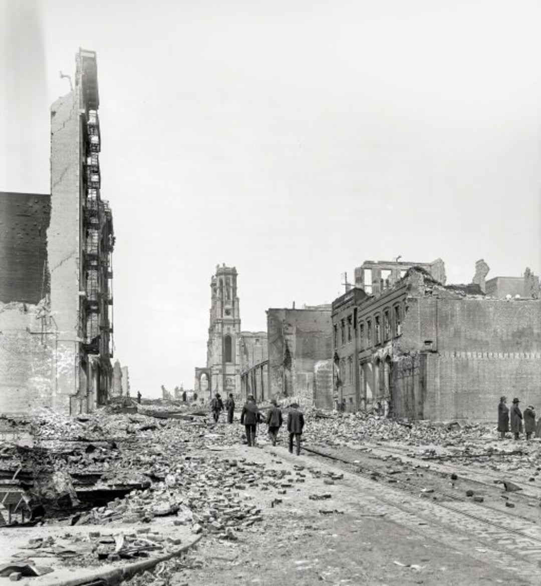 People walk along a street filled with rubble and debris, surrounded by severely damaged and partially collapsed buildings, suggesting the aftermath of a major disaster or conflict.