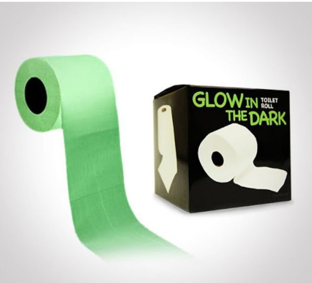 A light green toilet paper roll is shown next to its black packaging box labeled "Glow in the Dark Toilet Roll" in bold white and neon green text.
