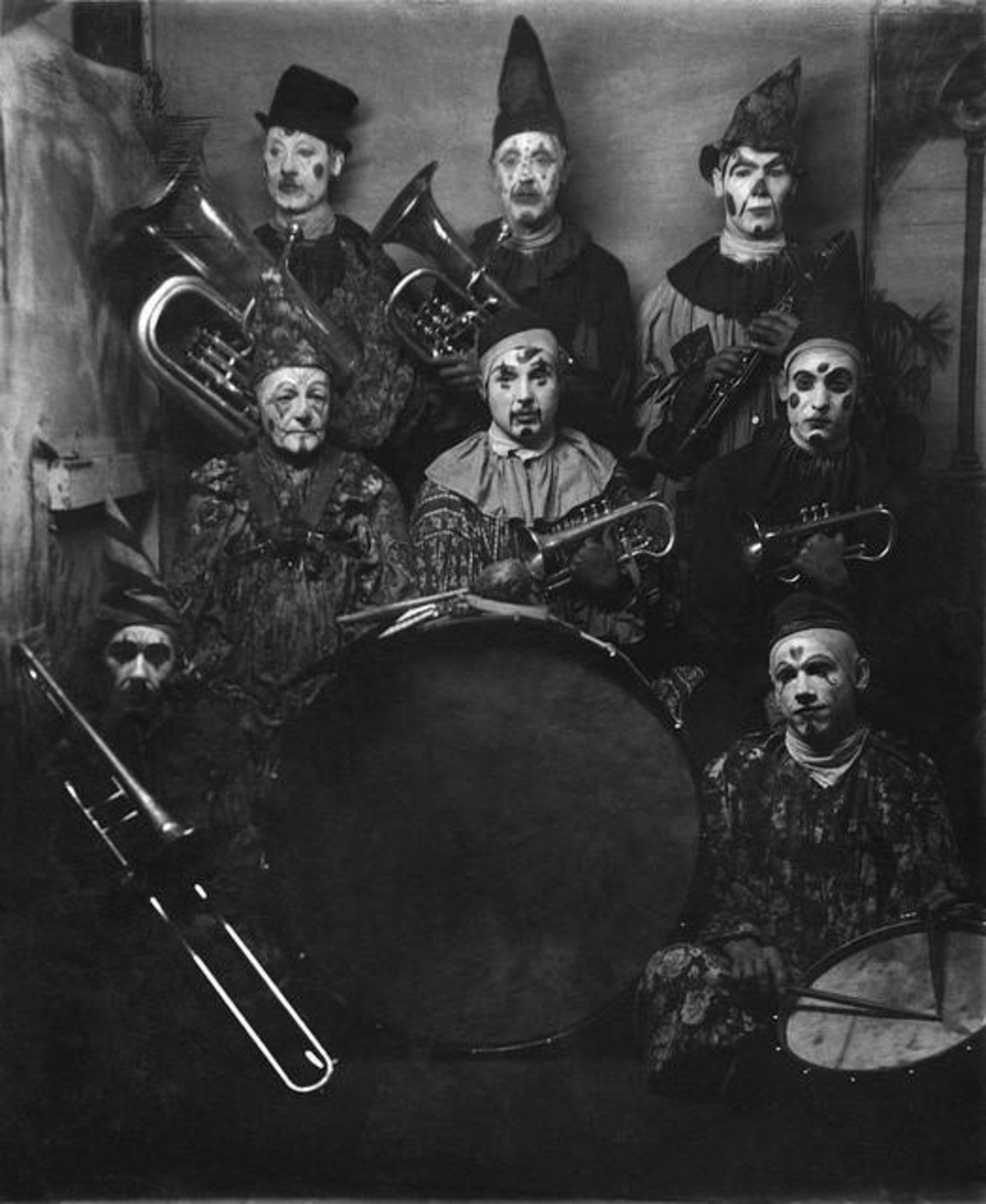 A black and white photo of eight clowns in costume and makeup, each holding musical instruments including brass horns, a drum, and a trombone, posing for a group portrait against a plain backdrop.