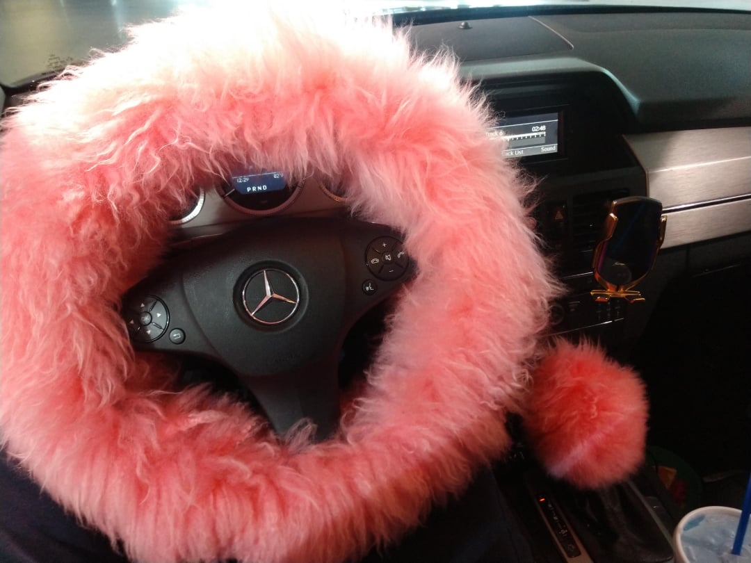 A close-up of a car interior shows a pink fluffy steering wheel cover and a matching fluffy gear shift cover. The car is a Mercedes-Benz, and sunglasses hang near the dashboard.