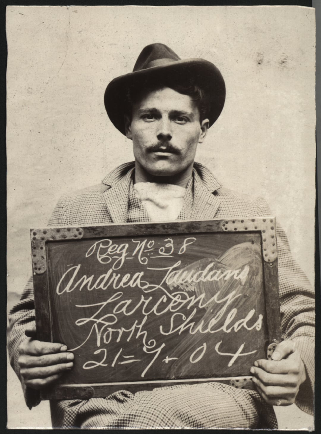A man with a mustache and hat sits, holding a sign reading: "Reg No 38 Andrea Laudana Larcon North Shields 21-9-04." The photo is black and white and appears old-fashioned.