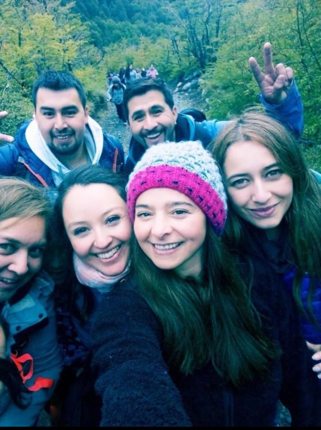 A group of six smiling friends, bundled in warm clothes, take a selfie outdoors on a wooded trail. Other people can be seen walking in the background among green trees.