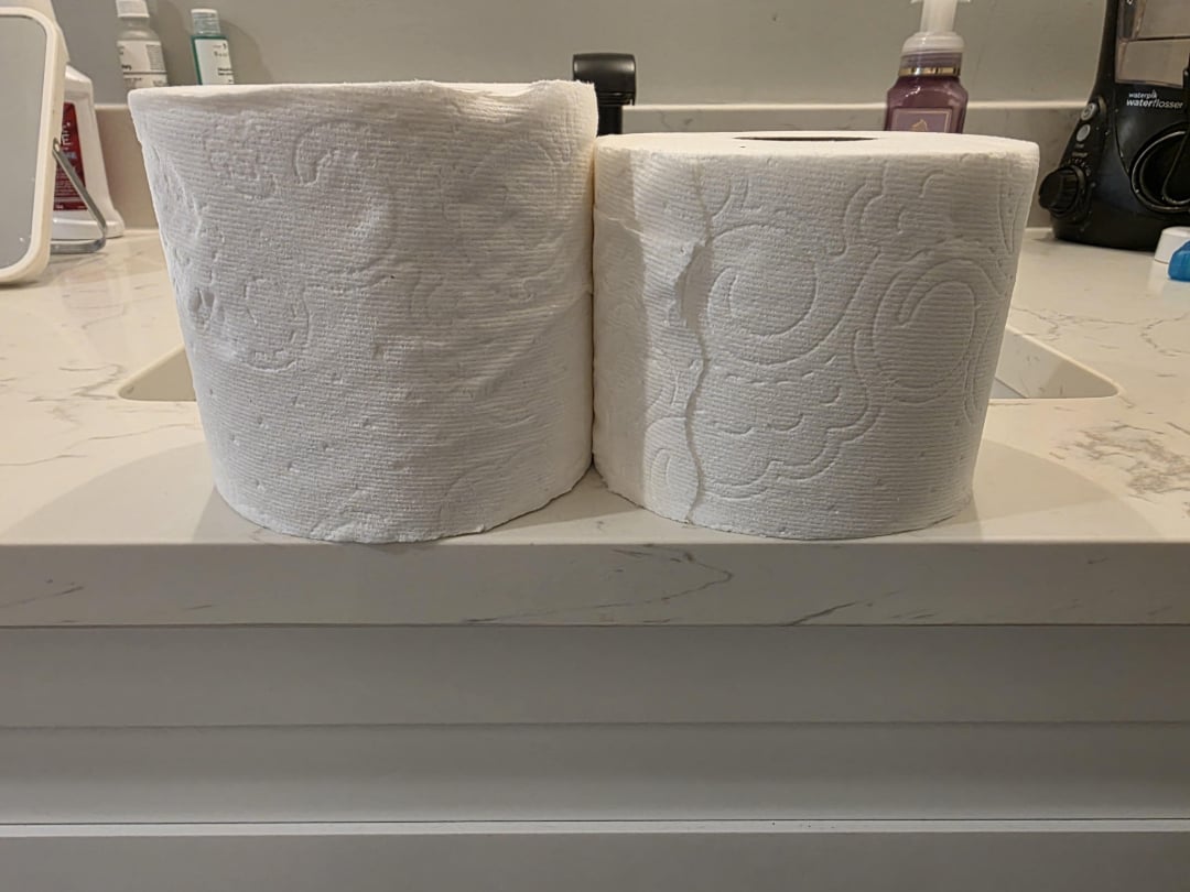 Two rolls of toilet paper side by side on a bathroom counter, with one roll noticeably larger than the other. Bathroom items and soap dispenser are visible in the background.