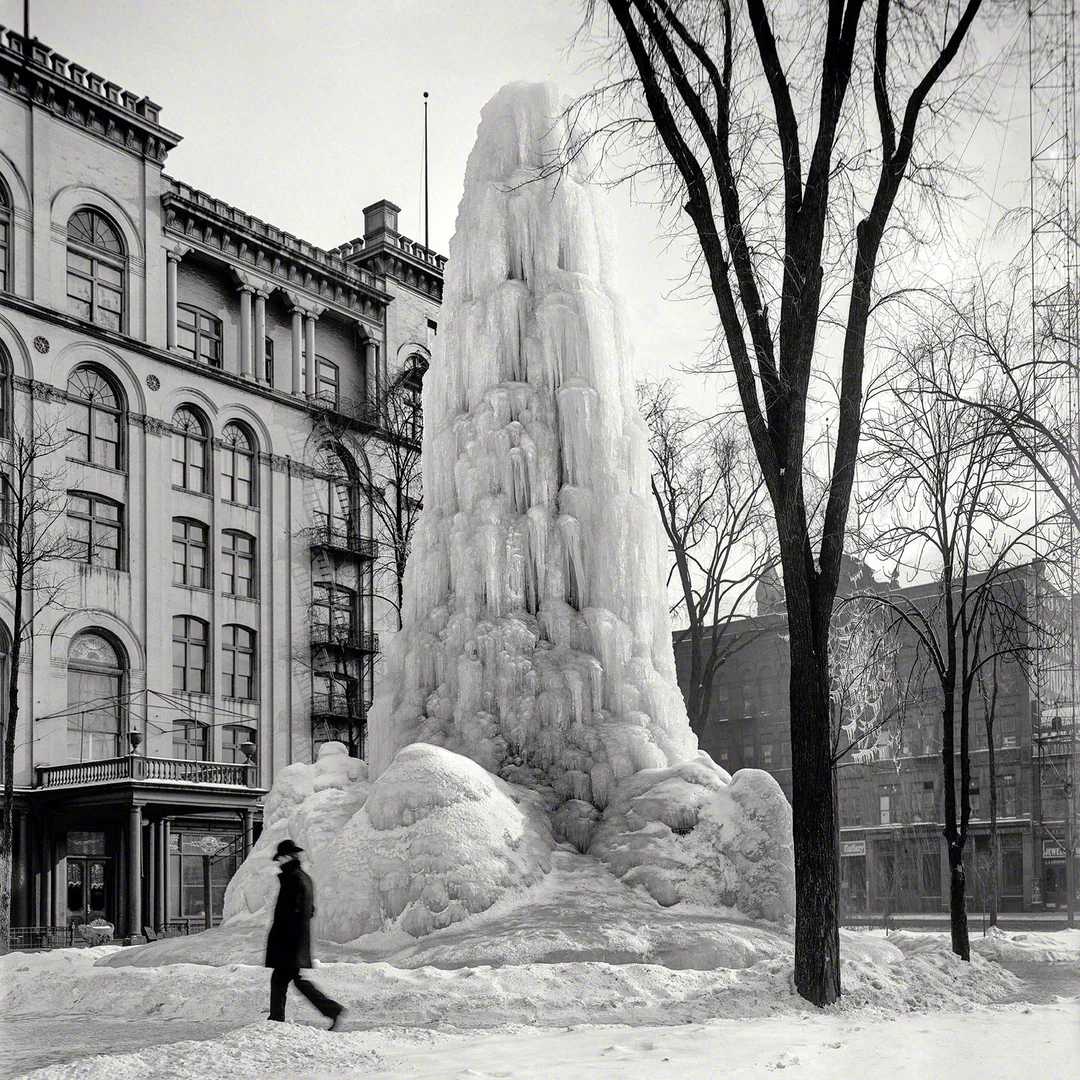 A person in a long coat and hat walks past a large, towering ice formation in a snowy city park, surrounded by bare trees and historic buildings in the background.