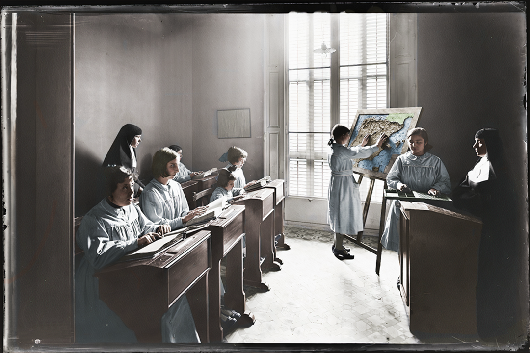 A vintage classroom scene showing young girls in blue uniforms seated at wooden desks, with two nuns supervising. One girl stands at an easel pointing to a map, sunlight streaming through tall windows behind her.