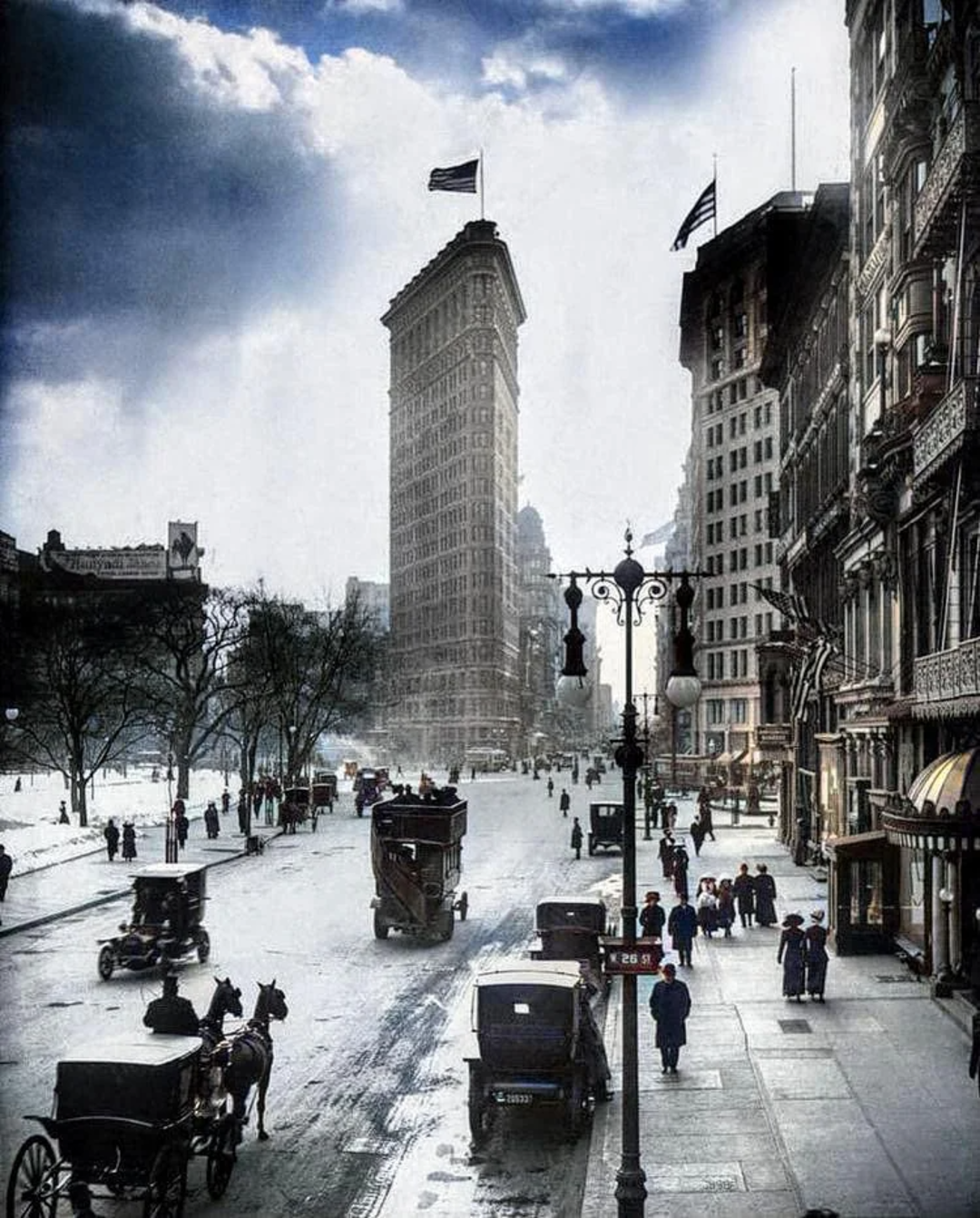 A bustling early 20th century city street scene with horse-drawn carriages, vintage cars, and people walking. The Flatiron Building is prominently visible in the background under a partly cloudy sky.