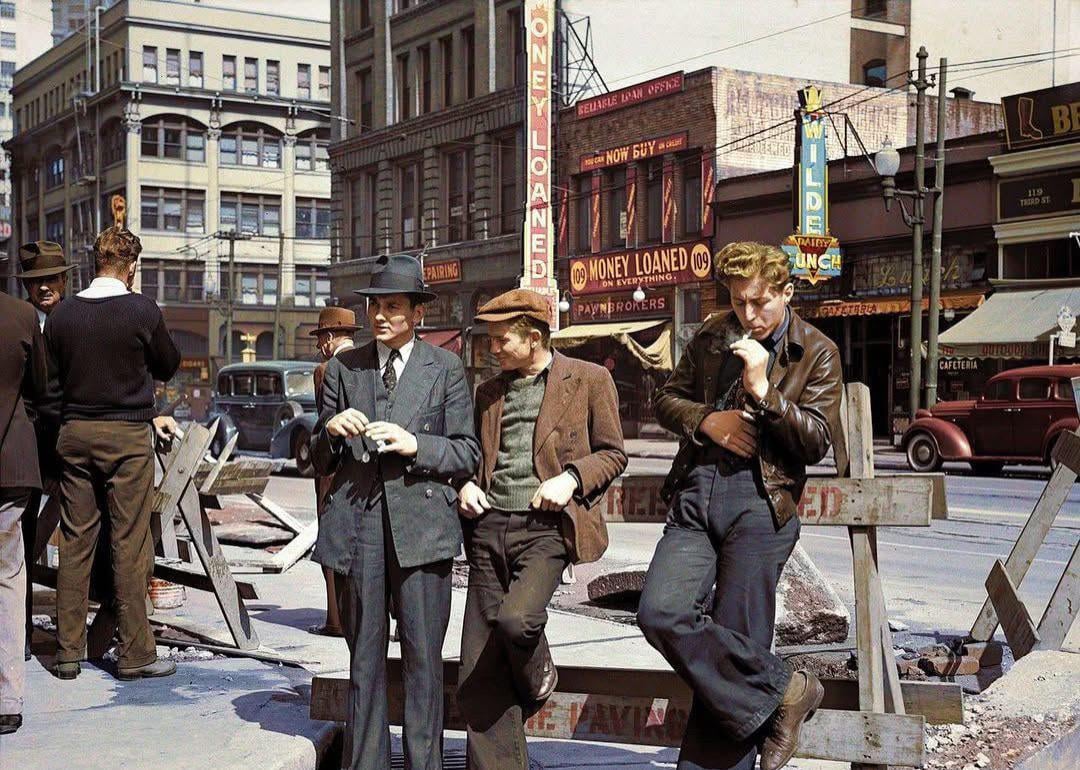 Three men in 1930s-style clothing stand by a street barricade in a city, with businesses and vintage cars in the background. Signs read "MONEY LOANED." Urban construction is visible around them.