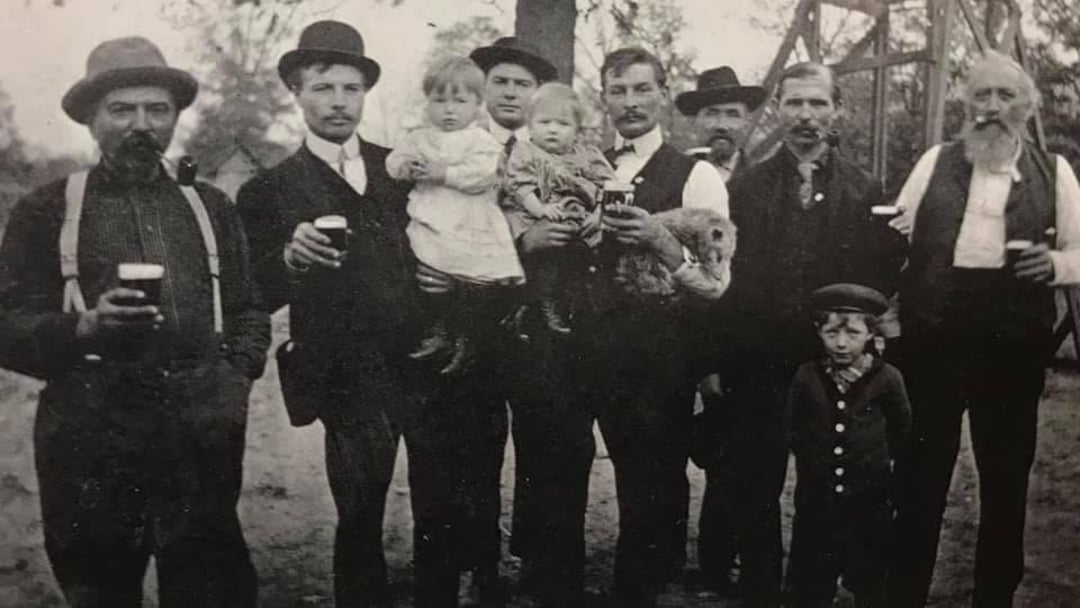 A vintage black-and-white photo shows a group of men standing outdoors, some holding children or drinks, dressed in old-fashioned clothing and hats. Two small children stand in front, and trees are visible in the background.