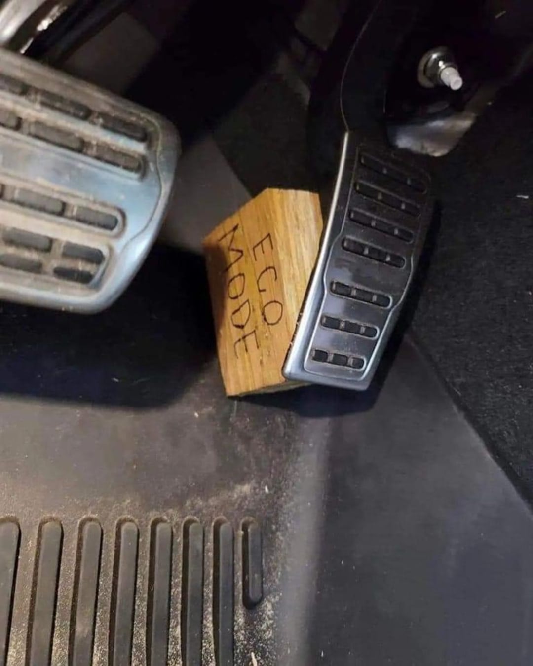 A wooden block labeled "EGO MODE" is wedged under a car’s accelerator pedal, preventing it from being fully pressed down. The block is positioned between the gas pedal and the car’s floor.