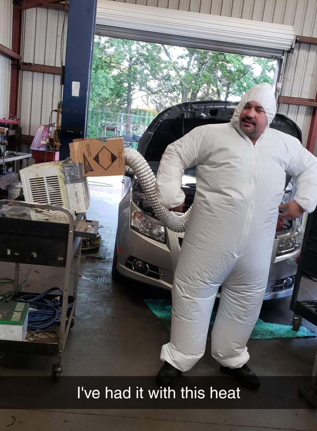 A man in an inflated white suit stands in a garage, with a flexible duct from a window air conditioner running into his suit. A car with its hood up is behind him. Text at the bottom reads, "I've had it with this heat.