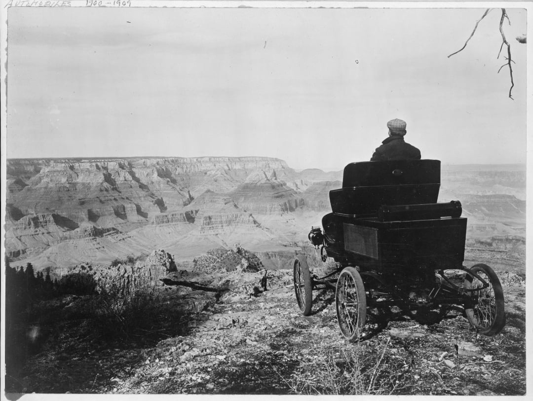 A person sits in an early automobile at the edge of the Grand Canyon, gazing out over the vast, rugged landscape under a clear sky. The scene evokes a sense of solitude and exploration.