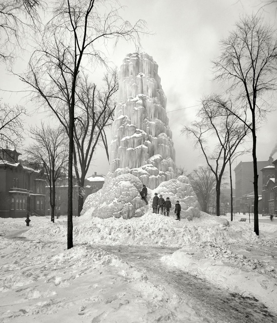 A group of people stand near a tall, layered ice tower in a snowy park surrounded by bare trees and buildings on a winter day. Paths are visible in the snow leading to the ice structure.