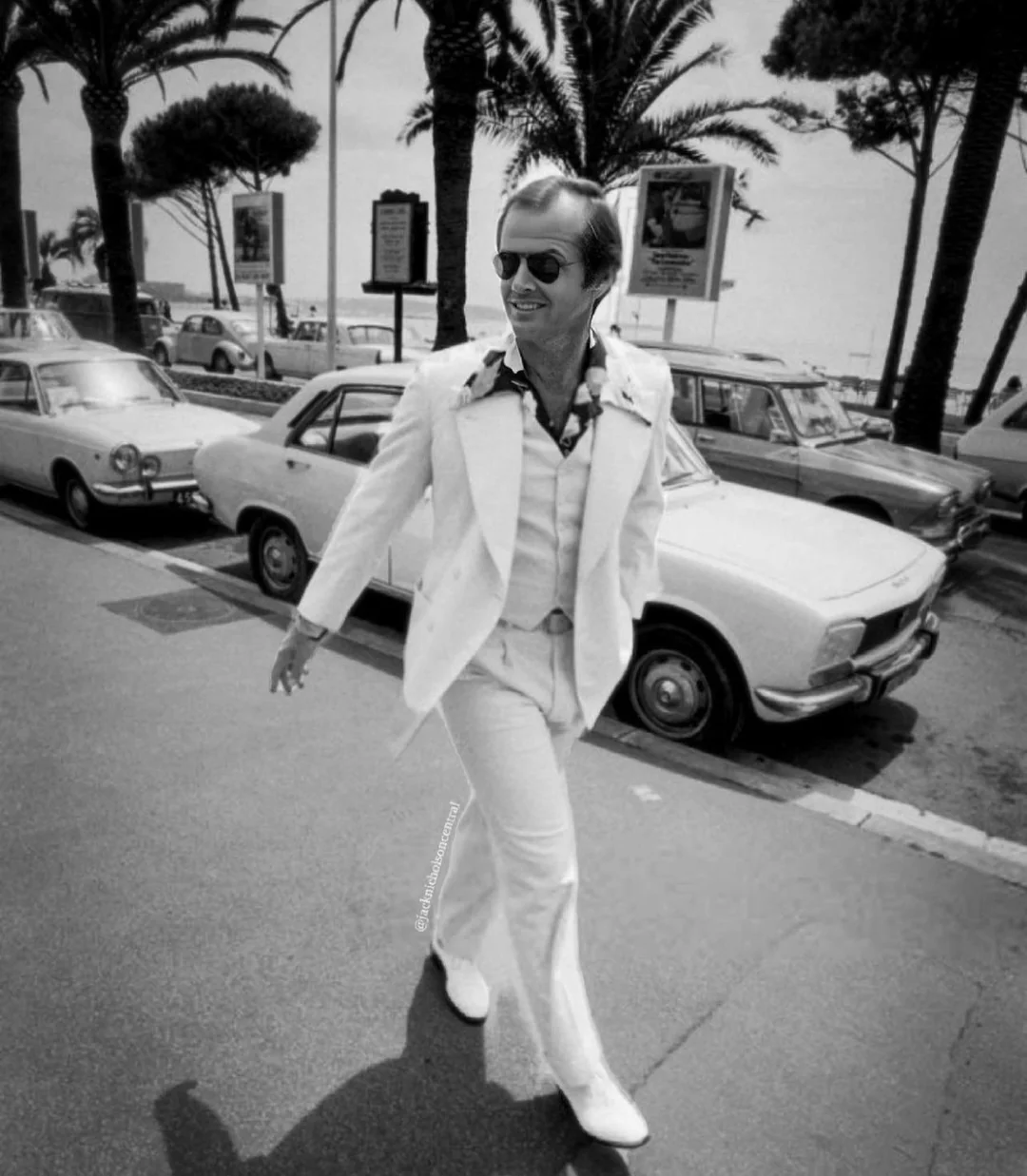 A smiling man in a white suit, patterned shirt, and sunglasses walks confidently on a street lined with classic cars and palm trees on a sunny day. The scene appears vintage, likely from the 1970s.