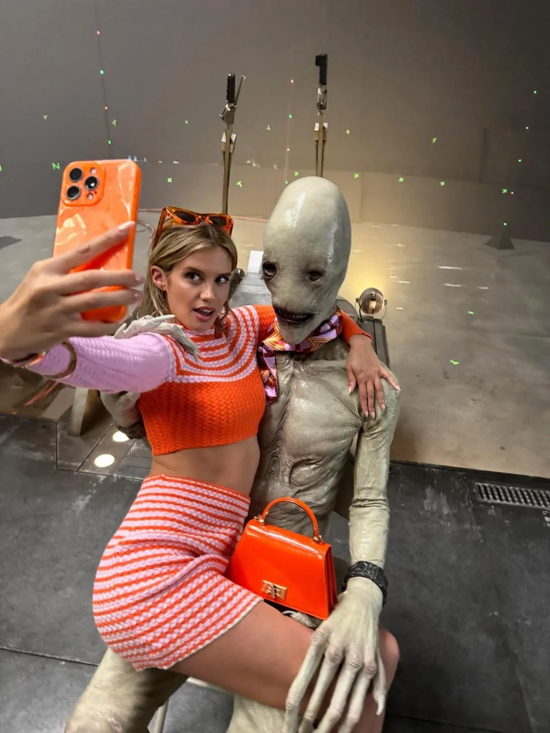 A woman in a pink and orange outfit takes a selfie while sitting on the lap of a tall humanoid creature with pale, textured skin and a wide mouth. She holds an orange phone and purse, and the scene appears to be on a film set.