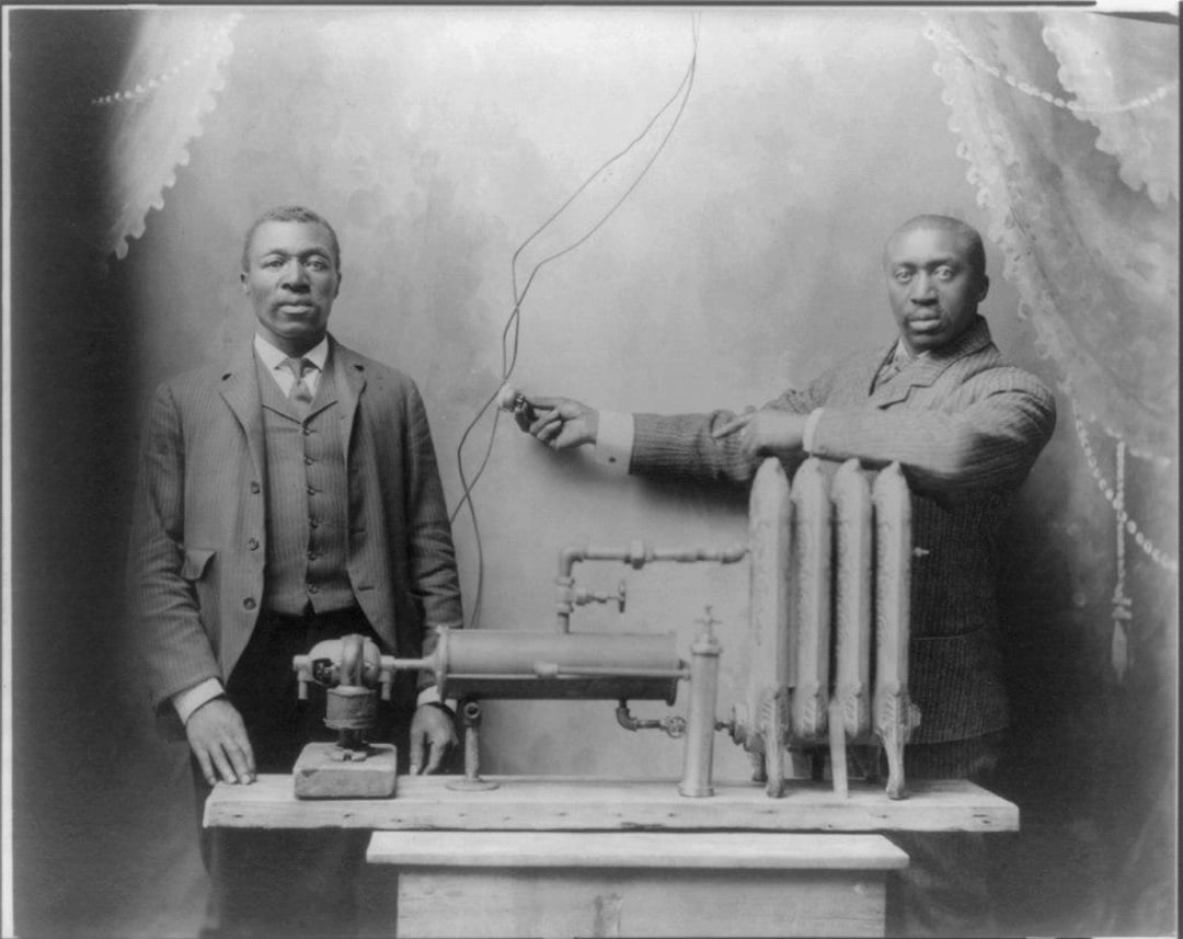 Two Black men in suits stand beside an early mechanical device on a table. One man holds a wire leading from the machine, while the other stands with his hands at his sides. The backdrop has draped curtains.